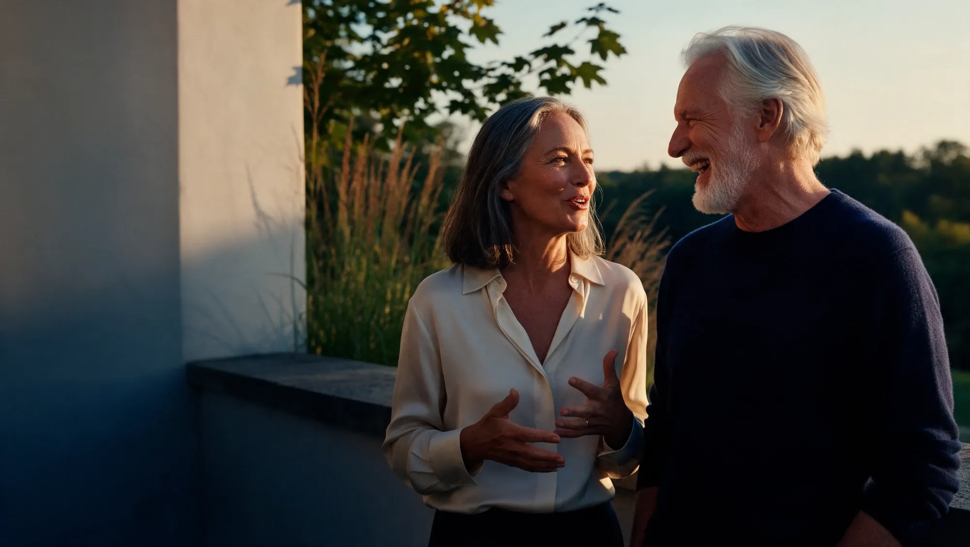 A woman and a man in their mid-fifties talking on a stone terrace at golden hour, she is mid-sentence and he is laughing