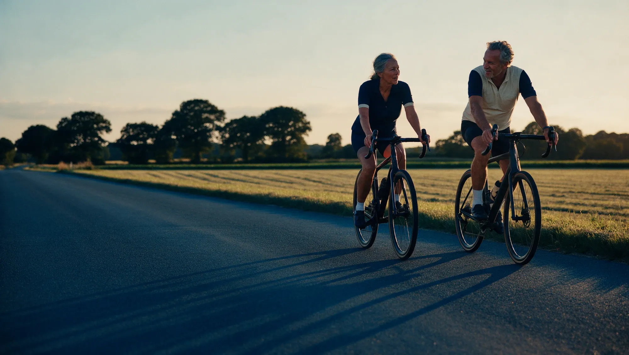 A man and a woman in their late fifties cycling side by side on a quiet country road at golden hour